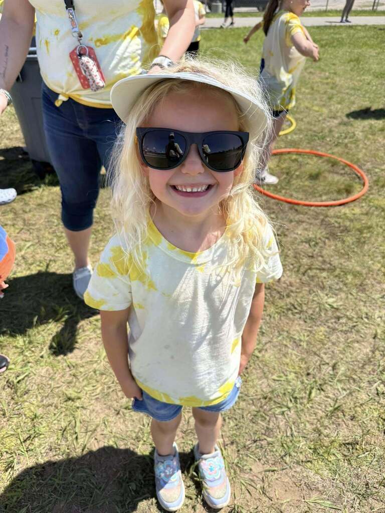 Child with blonde hair, yellow tie-dye shirt, sunglasses, and a white hat stands on a grassy field.