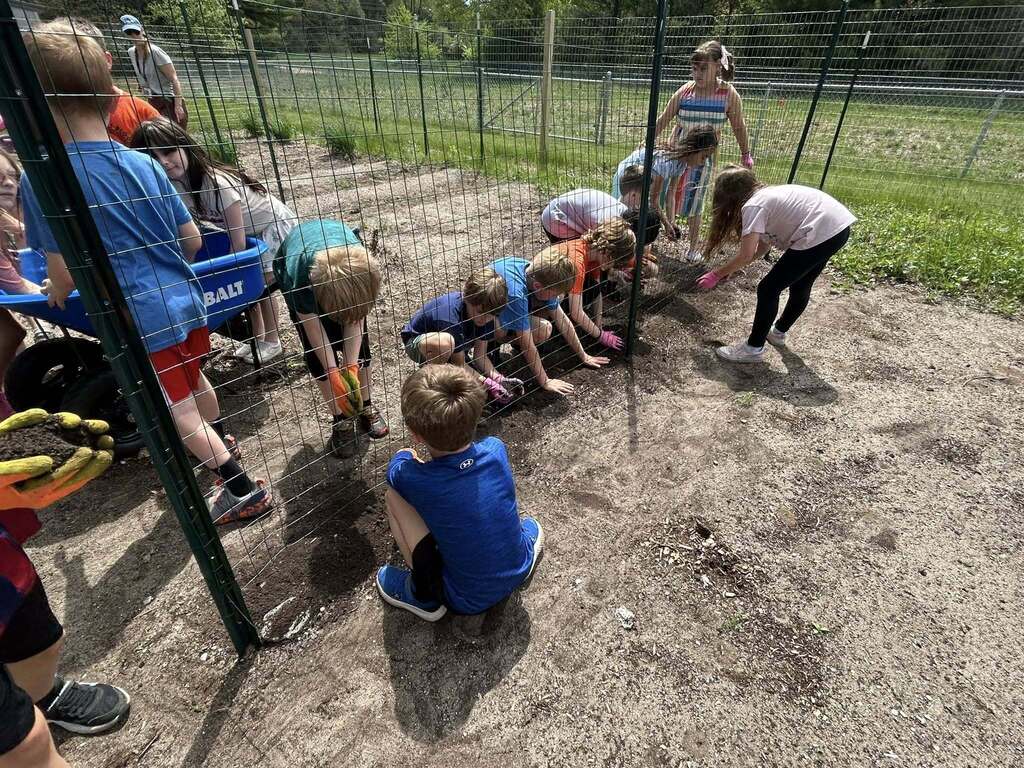 Children and adults in a dirt area behind a fence, engaging in outdoor activities, some seated and others standing.