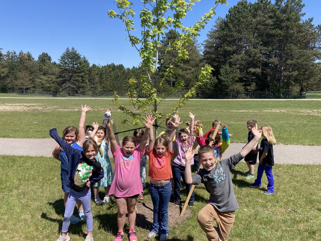 A group of children in a park pose around a young tree with raised hands. Trees in the background.