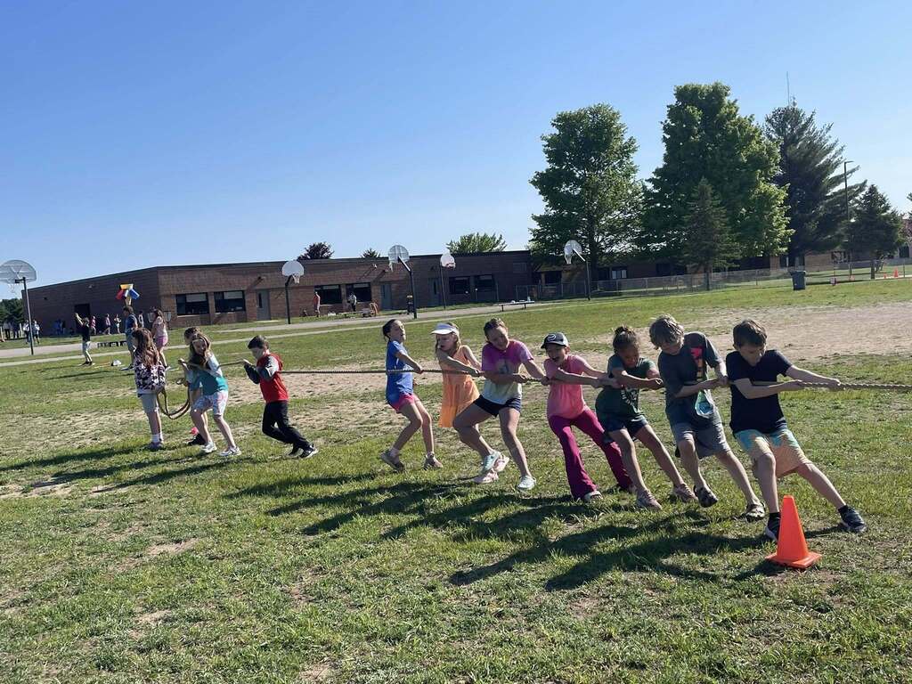 Group of children pulling on ropes during a tug-of-war in a grassy field. Trees and a building are in the background.
