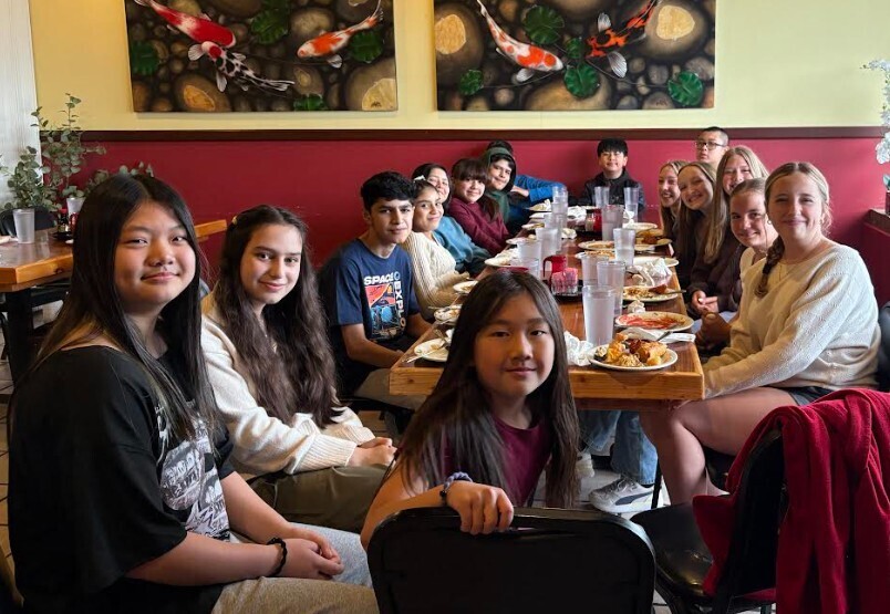 A group of people sit around a long table in a restaurant, smiling, with fish paintings on the wall.