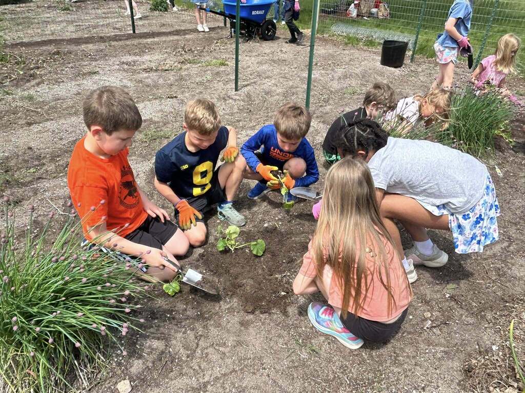 Children plant seeds in a garden area. Some wear gloves and hold tools. Background shows a fenced garden with plants.