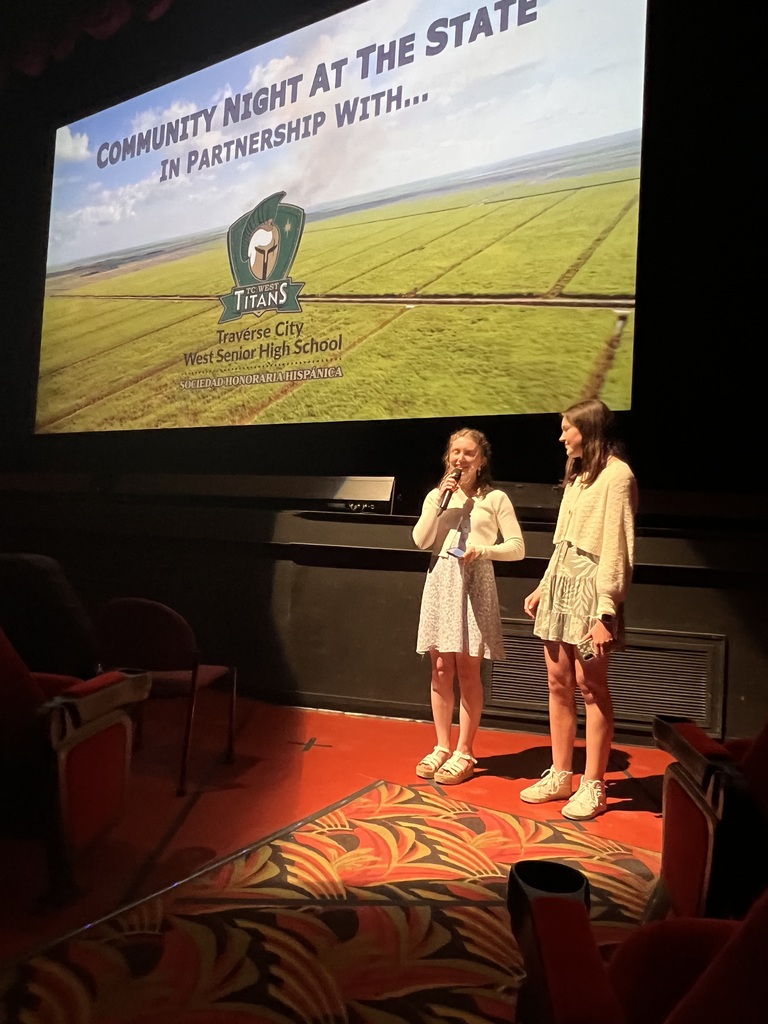 Two women stand on stage, one holding a microphone, with a large screen behind them displaying a field.