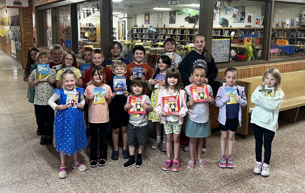 Group of children in a library, holding books, with a library background and a bench.