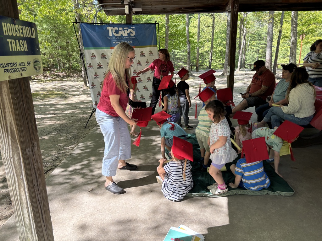 Woman in red shirt holds red graduation caps for children kneeling on a rug. Banner with "TCAPS" and trees in background.
