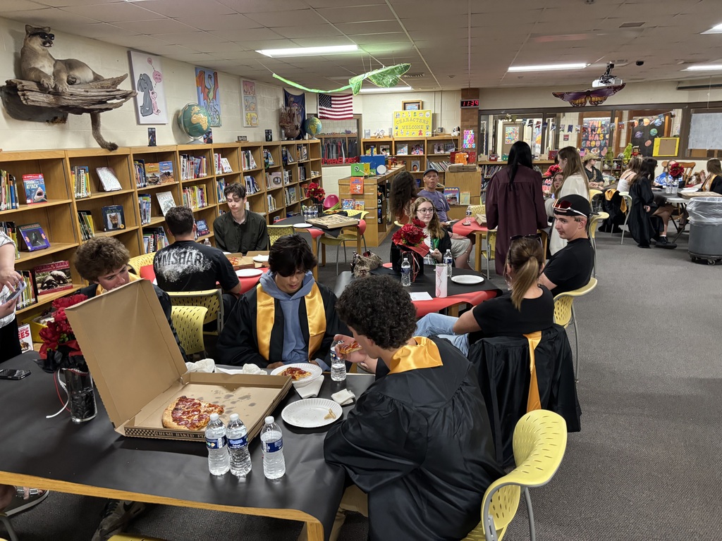 People in robes sit around tables in a library, eating pizza and using plates. Books on shelves and decorations are in the background.