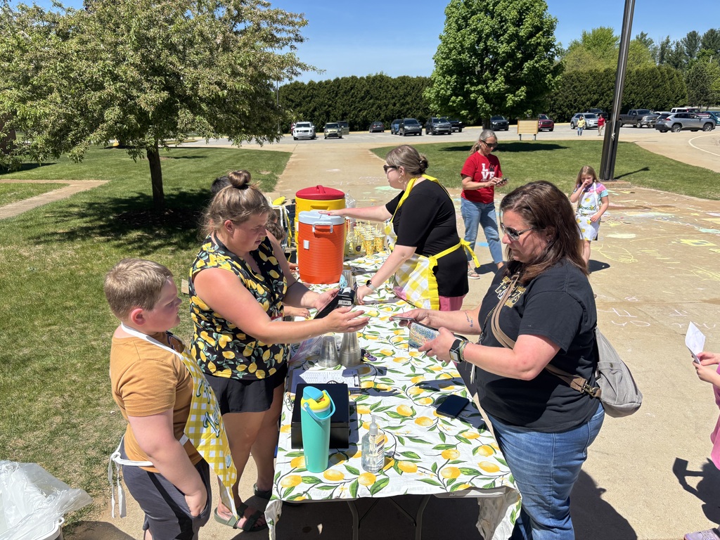 Several people stand around a table with a yellow and white tablecloth, preparing something. Trees and a grassy field are in the background.