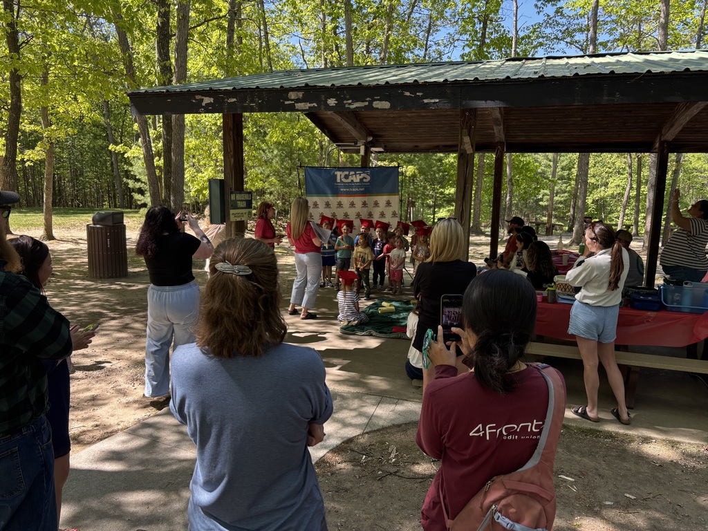 People gathered in a wooded area, some taking photos, beneath a shelter with a banner.