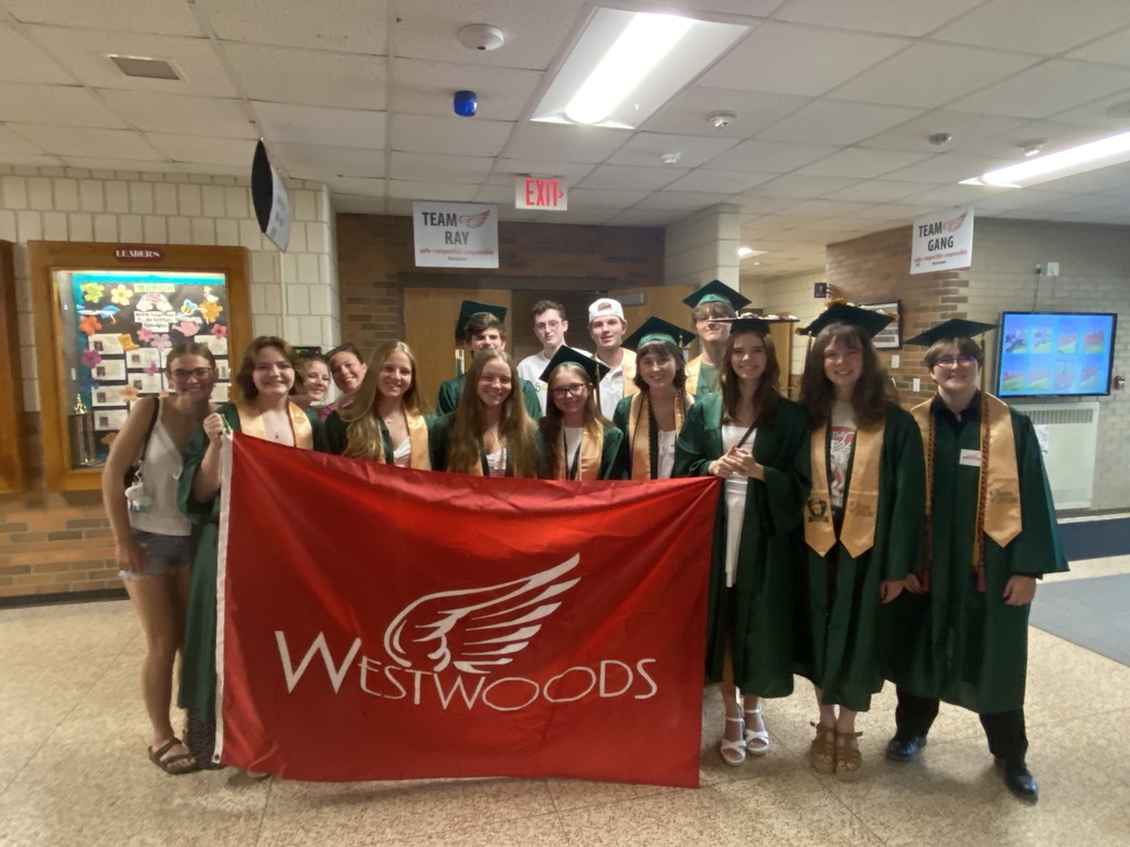A group of people in graduation attire standing in a hallway. They hold a red banner that reads Westwoods.