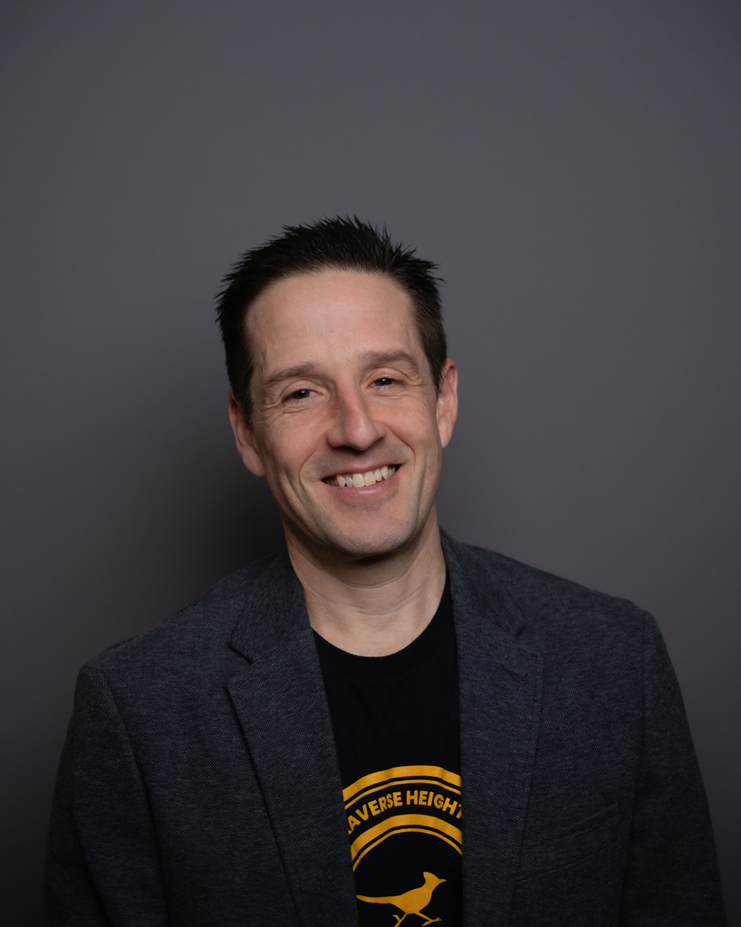 A smiling man with short hair stands against a dark background. He wears a black shirt with a yellow graphic and a gray blazer.