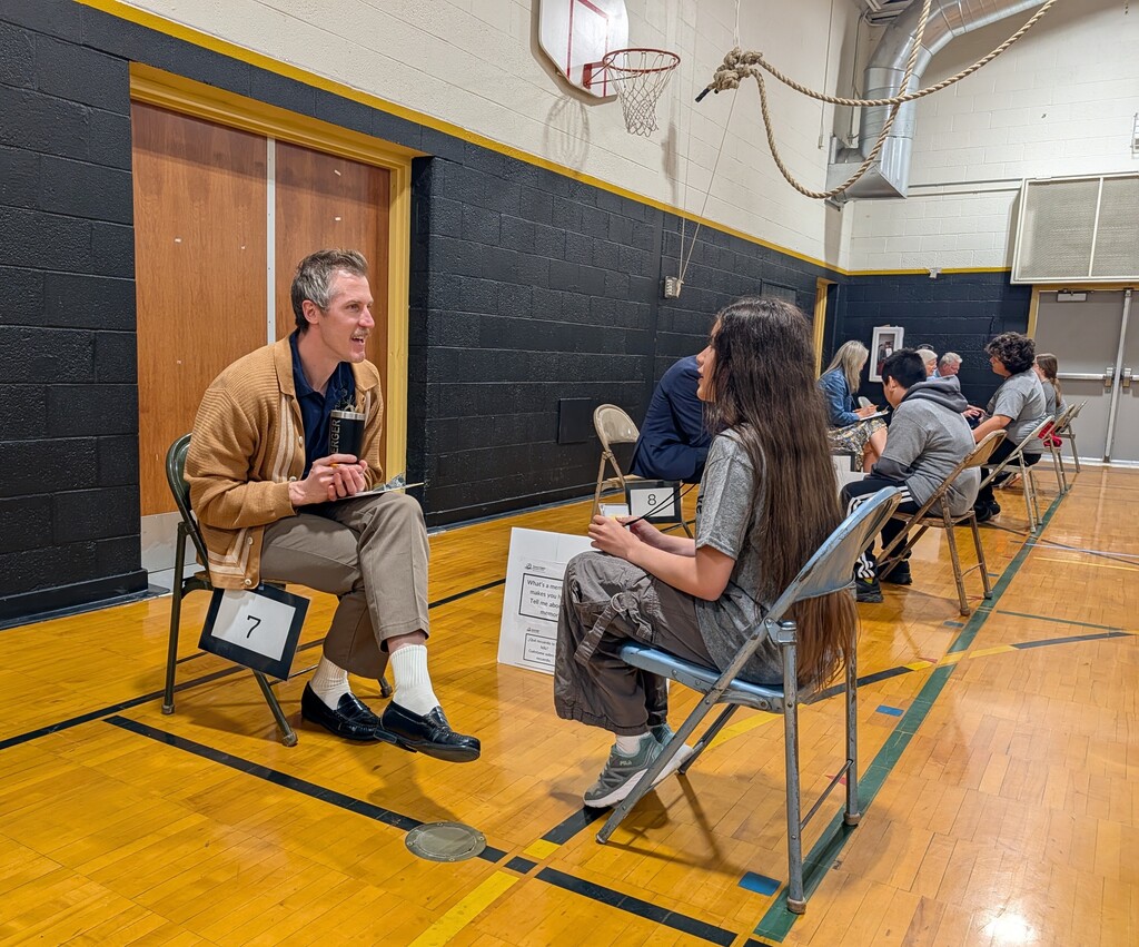 Two people sit in chairs inside a gym, labeled with numbers. A basketball hoop is in the background.