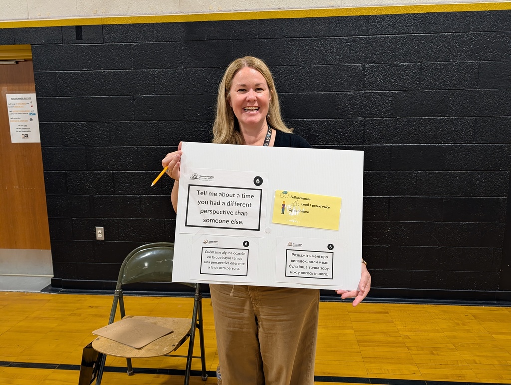 Woman in a gym holding a board with text and sticky notes. A chair and wooden wall in the background.