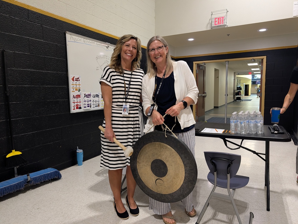 Two women in gym, one holding a gong, with a table and chair nearby, a whiteboard on the wall.
