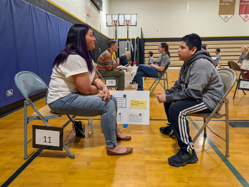 People sit in chairs with numbered signs, conversing in an indoor gym setting. The floor has wooden panels and markings.