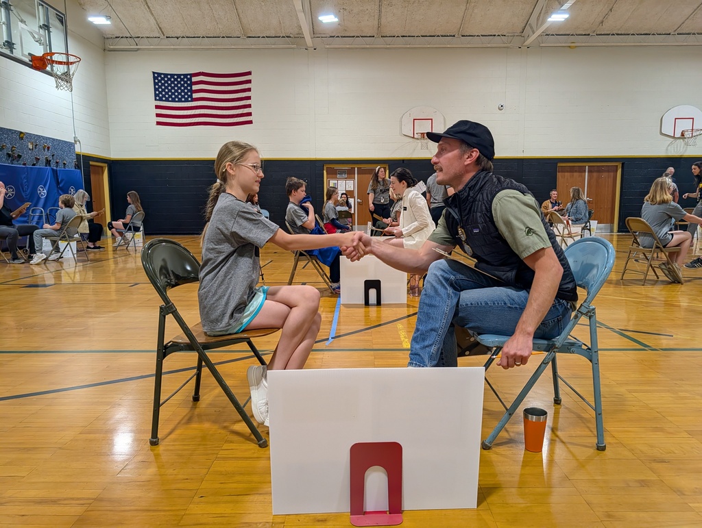 A young girl and a man sit in chairs shaking hands in a basketball court. Other people are sitting at tables.