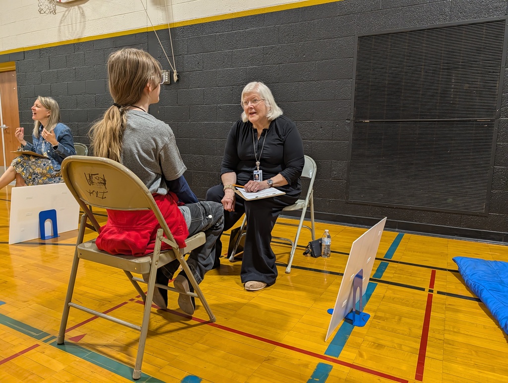 Two women in chairs, one holding a clipboard, converse in a gym with a wooden floor and black walls.