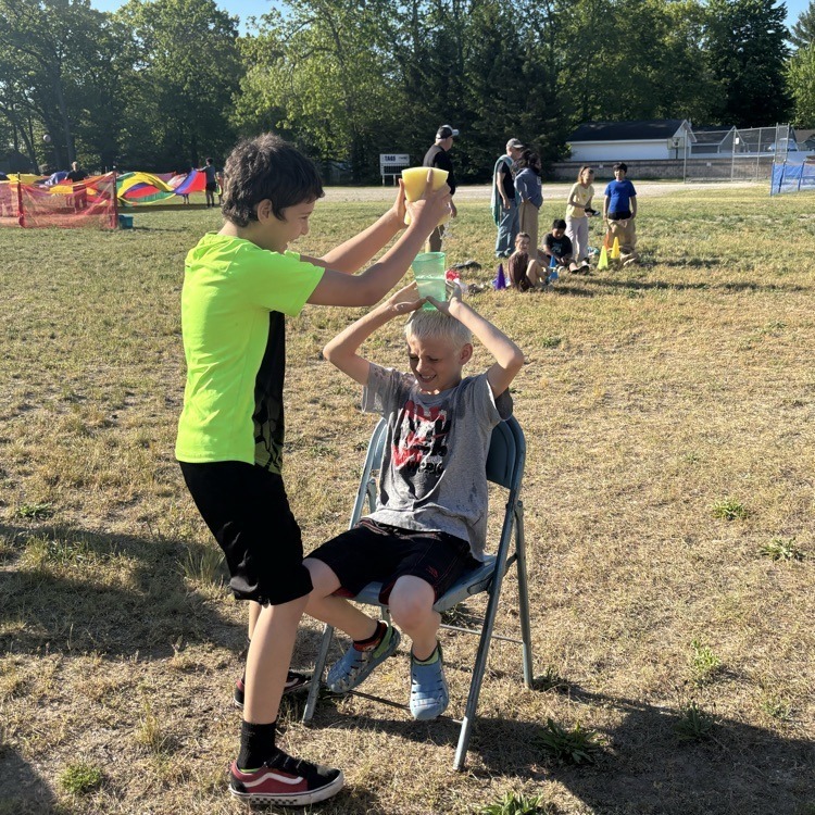 A boy pours water from a yellow cup over another boy's head. They sit in an outdoor field with other kids.