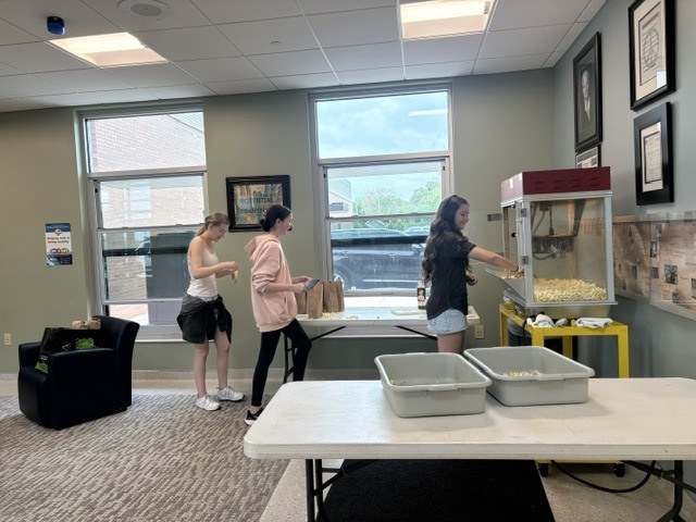 Three female students in a room, one by a popcorn machine, another by a table, and a third by a window.