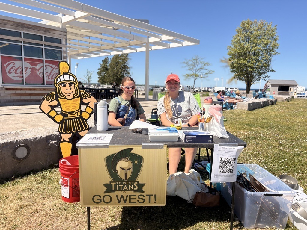 Two individuals are sitting at a table outside, with items like a helmet and a sign that says Titans Go West.