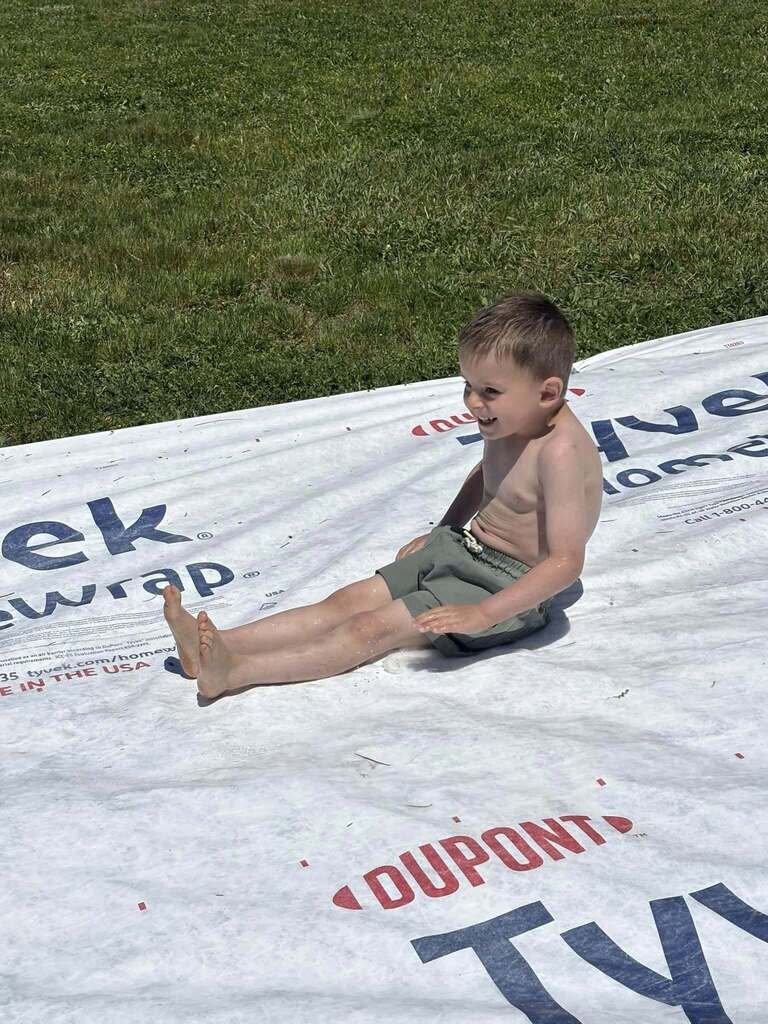 A boy sits on a tarp with "Dupont" and "Tyvek Wrap" written on it, against a grassy background.