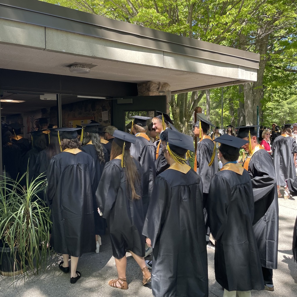 Graduates in black caps and gowns stand in a line, some with caps on, outside a building.