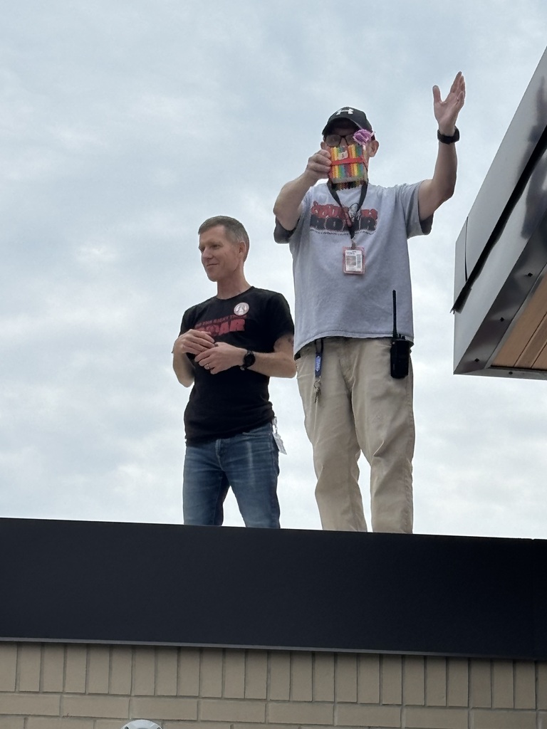 Two individuals standing on a rooftop, one holding a camera and the other waving with one hand.