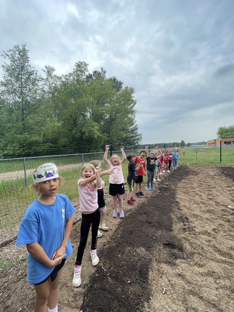 Kids stand in a line in front of a fence. Some have hands raised. A field with dirt is behind them.