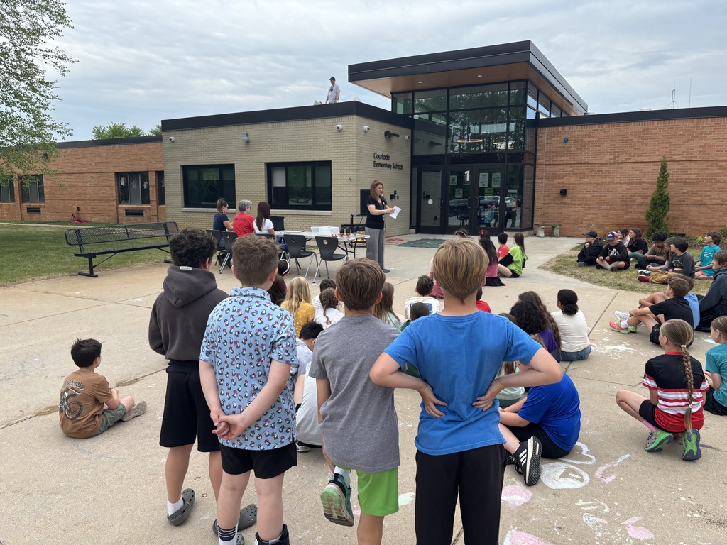 Kids of different ages are gathered outside a building, listening to a person standing in front of them.