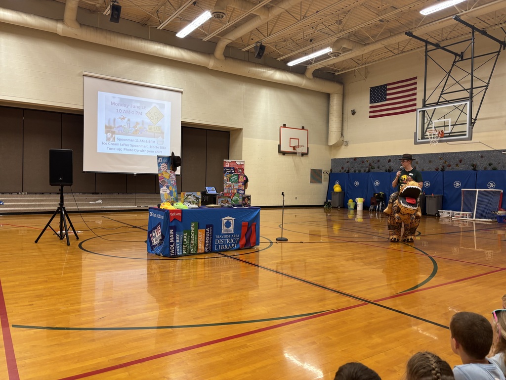 Indoor gymnasium with a projector screen, a mascot, and a table with books. Children seated in the background.