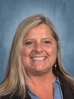 Woman with long, blonde hair wearing a blue shirt and black jacket smiles at camera.