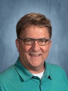 A man with short brown hair wearing glasses and a green polo, smiling against a blue background.