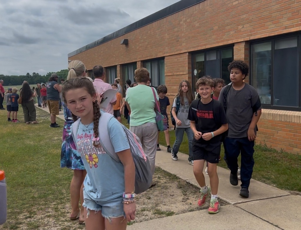 A line of children and adults walk on a sidewalk in front of a brick building.