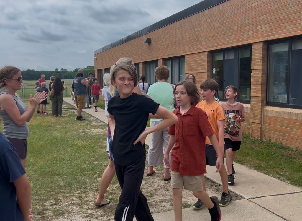 People, including children, walk along a sidewalk in front of a building. Some wear casual clothing.