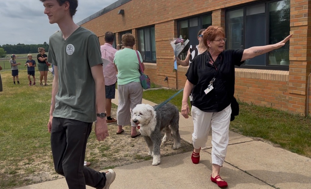 Several people walk along a sidewalk in front of a building, including a woman with a dog.