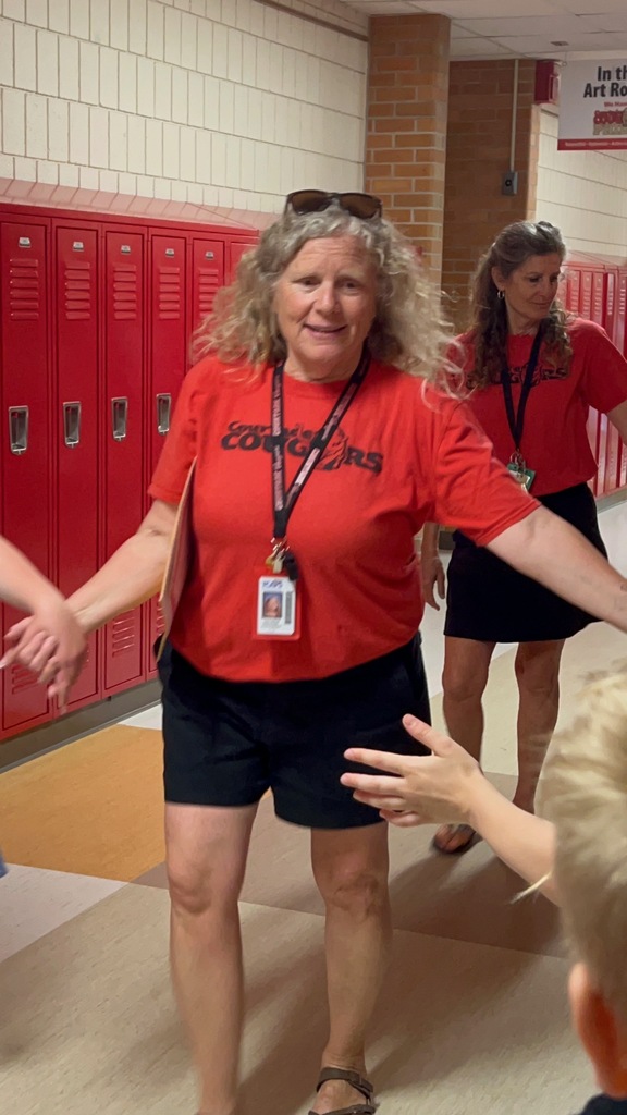 A woman in a red shirt welcomes a student with a handshake in a hallway with red lockers and beige carpet.