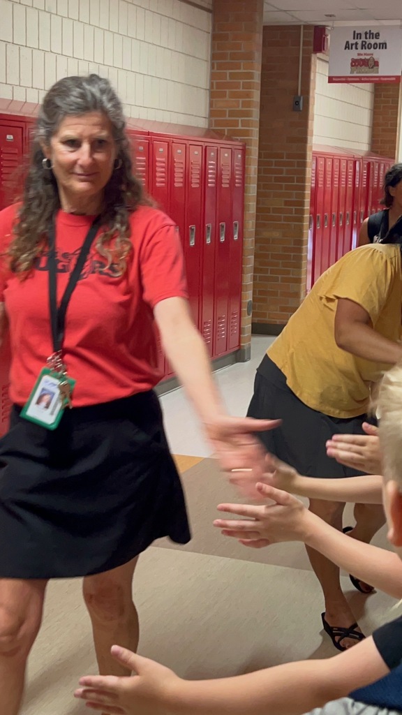 A woman in a red shirt and black skirt dances with two children in front of red lockers.