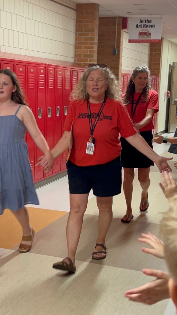 A hallway with red lockers. A woman in a red shirt stands with two other women. People reaching out to greet them.