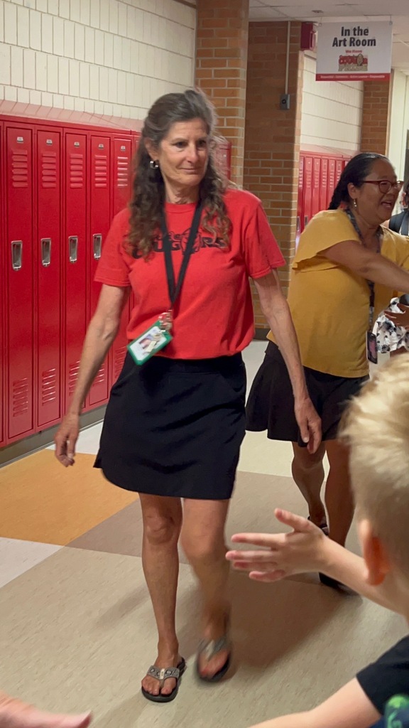 A woman in red walks in a school hallway with a boy extending a hand. A sign reading "In the Art Room" hangs above red lockers.