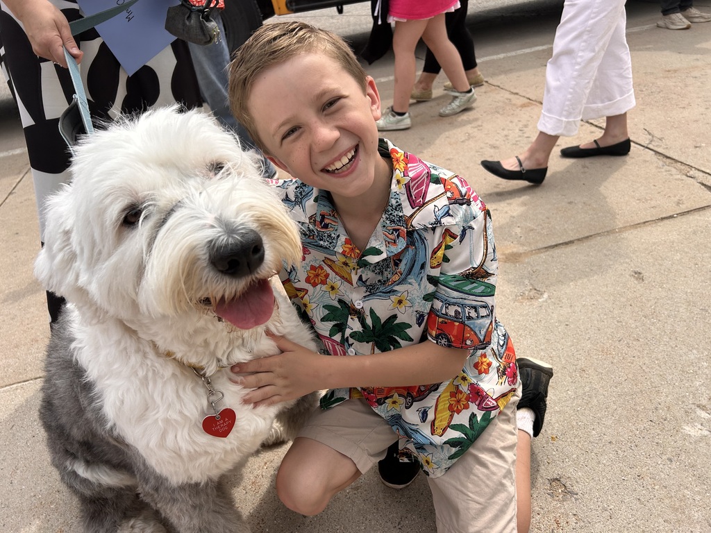 A smiling young boy with a Hawaiian shirt kneels next to a fluffy white dog. Behind them, several people stand on a sidewalk.