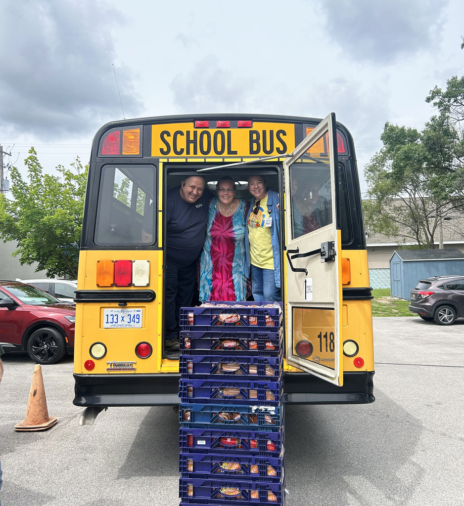 Three people stand in a school bus with crates stacked at the rear. Behind the bus is a parking lot with trees and a building.