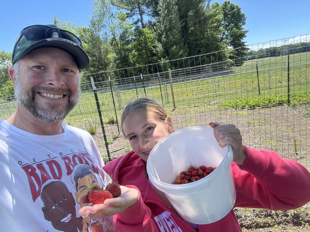 A man and a woman in a garden, smiling. They hold a bucket of strawberries and pose for a photo.