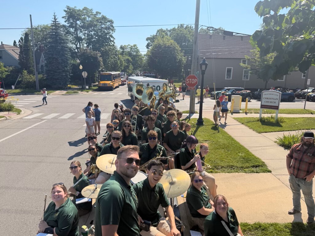 A marching band gathers in a street, with several members seated on chairs. They wear green shirts and play instruments.