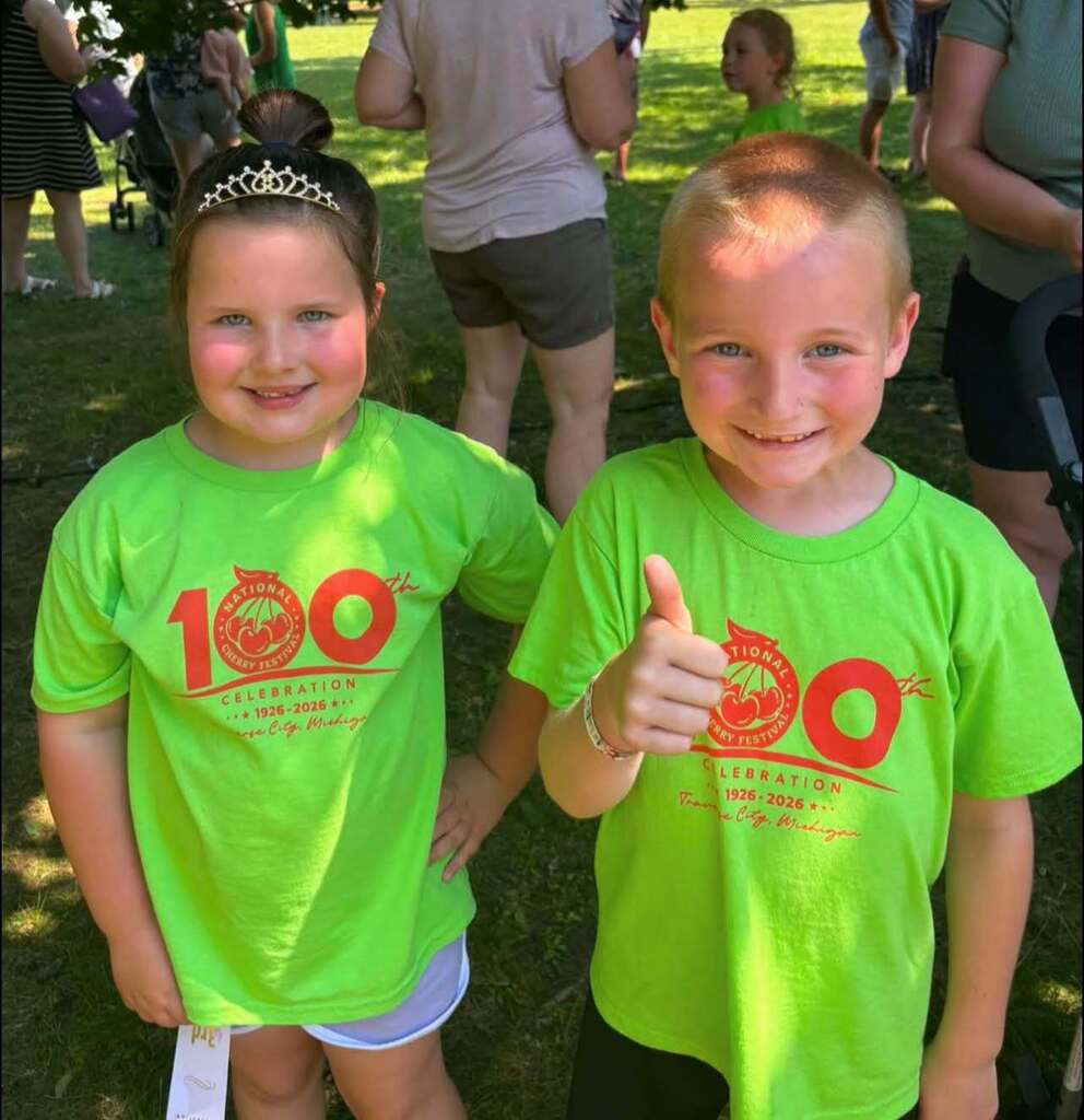 Two young kids wearing matching green t-shirts with red text pose for a photo. A blurry background shows people.