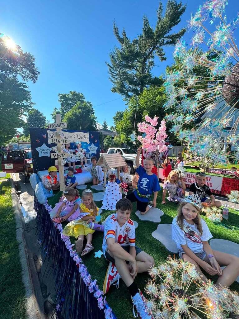 A float displays children in costumes; some sit on grass, others stand. Decorative elements include balloons and streamers. Trees and a building visible in the background.