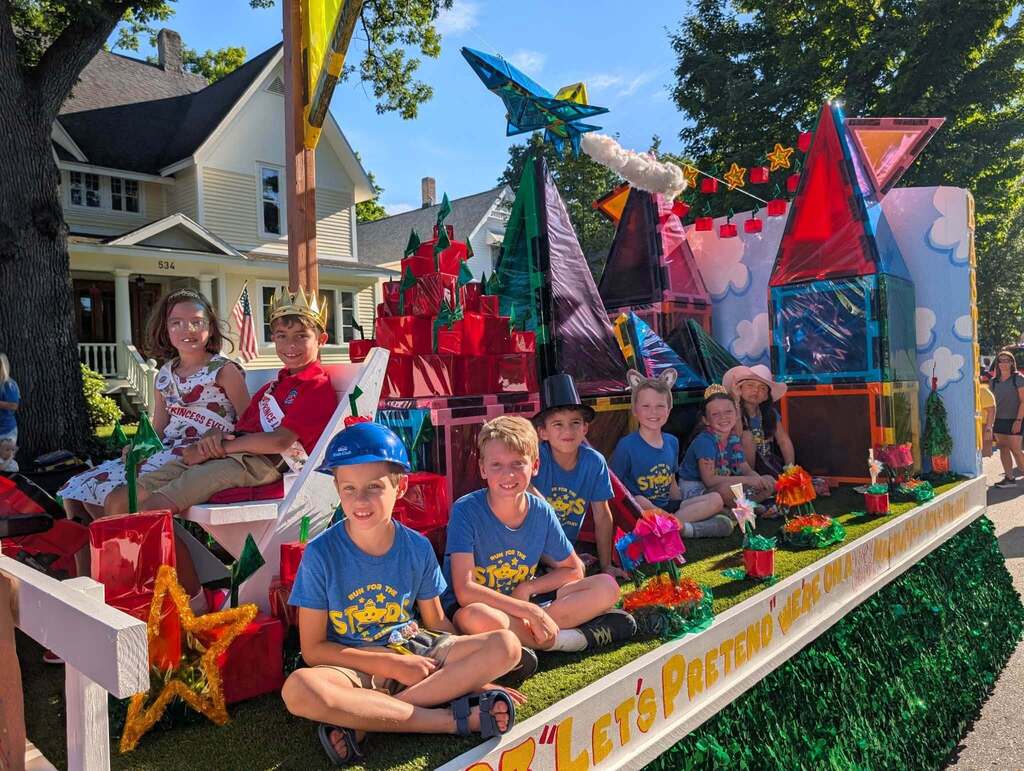 Children in matching T-shirts sit on a float decorated with colorful stars, windmills, and houses.