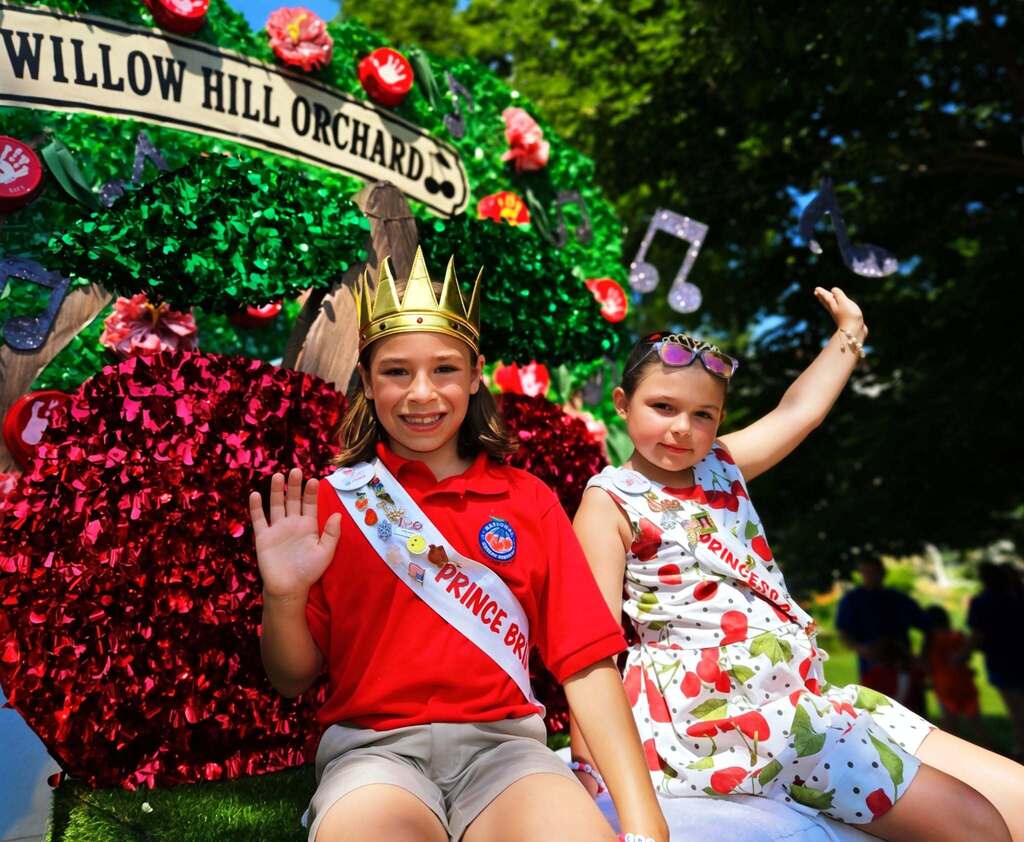 Two young girls in a decorated float, one wearing a crown and red sash, the other with a floral dress.