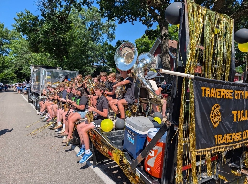 A marching band sits on a decorated float, playing instruments, with yellow balloons, a black banner, and a blue cooler.