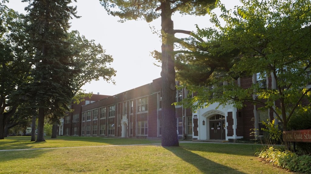 Bright sunlight over a grassy field and large trees in front of a brick building with arched doorways.