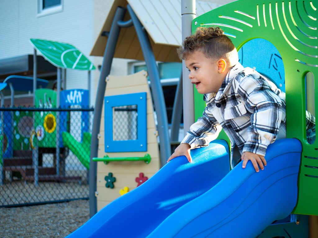 A young child plays on a blue slide in a playground with green and blue play equipment.