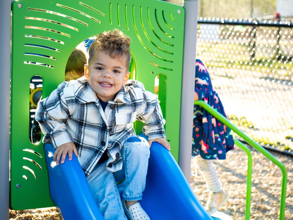 A child in a plaid shirt slides down a blue slide while smiling, with a green structure and a fence in the background.